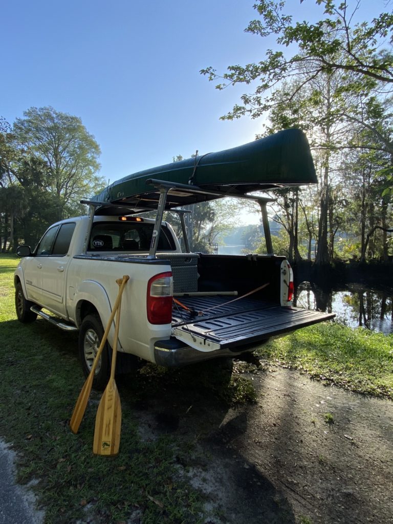 A white truck parked by the riverside with a new canoe on racks above the cab.