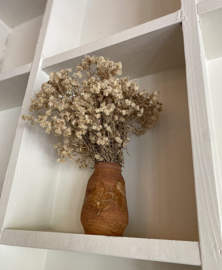 A bundle of dried flowers in a brown vase sitting on a white shelf